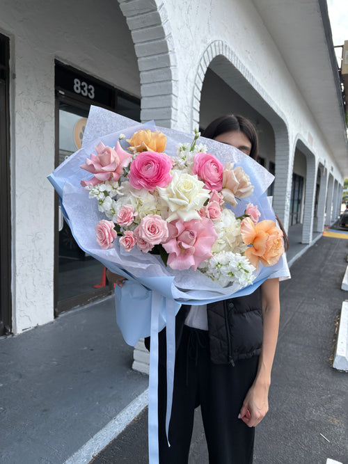 A person holding a bouquet of flowers with a blue wrap, consisting of pink, white, and orange flowers.
