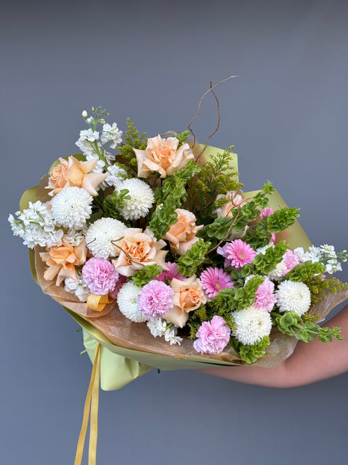 A hand holding a bouquet of mixed flowers including pink carnations, peach roses, white chrysanthemums, and green foliage, wrapped in brown paper and a pale yellow ribbon against a solid gray background.