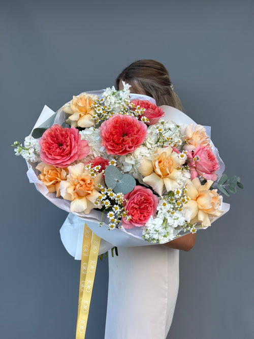A person in a white dress holds a large, round floral bouquet featuring vibrant pink and coral peonies, soft peach roses, white daisies, and green foliage, wrapped in white paper with a yellow ribbon, against a solid dark gray background.