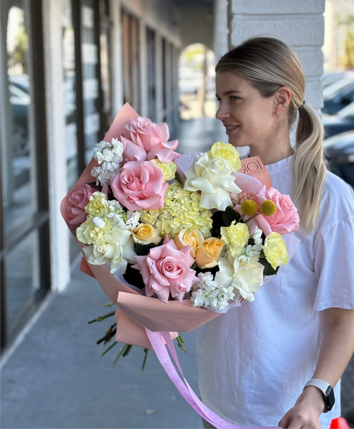 A person holding a bouquet of flowers, featuring pink roses and other mixed flowers in shades of yellow and green.