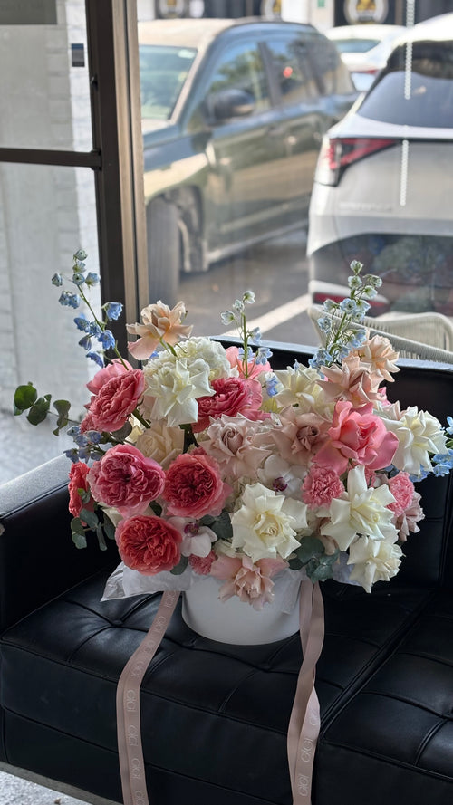 A floral arrangement with a variety of flowers including roses, in shades of pink, white, and green, tied with ribbons and placed in a white box.