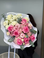 Bouquet of pink and white flowers wrapped in white paper held by a person against a neutral background.