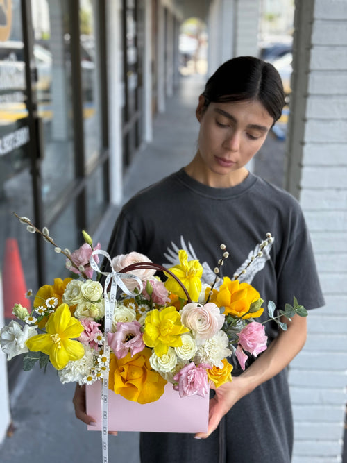 A person holding a bouquet of flowers with a variety of colors, predominantly yellow and pink, arranged in a box with a ribbon.