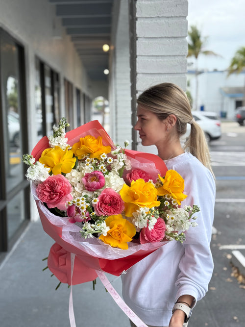 A person holding a bouquet of flowers which includes yellow, pink, and white flowers, with a red ribbon wrap.
