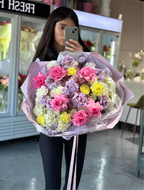 A person holding a large bouquet of flowers, featuring a variety of colors including pink, white, yellow, and purple, with a floral arrangement in the background.