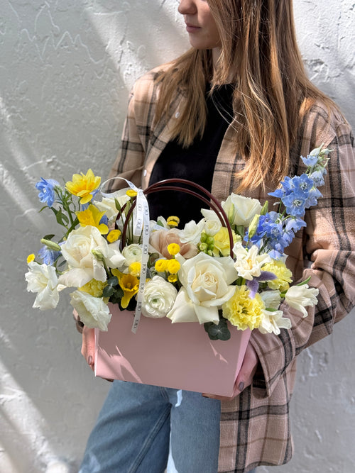 A person holding a pink floral handbag with a variety of flowers including white, yellow, and blue blooms.