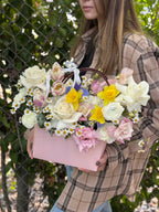 A person holding a pink floral arrangement with a variety of flowers including roses and other bloom types, in a handbag-style container.
