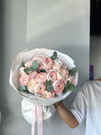 A person holding a bouquet of pink ranunculus flowers with green leaves, wrapped in a white and pink ribbon.