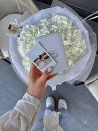Person holding a Fujifilm Instax camera with a large bouquet of white flowers in the background.
