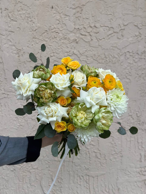 A bouquet of flowers including green roses, orange ranunculuses, white roses, and chrysanthemums, held against a neutral background.