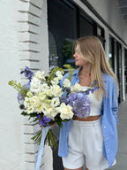 A person holding a bouquet of flowers featuring hydrangeas, lisianthuses, and roses in white, purple, and blue colors.