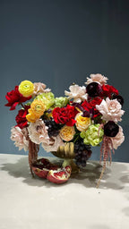 A floral arrangement featuring a mix of pink, red, and yellow flowers, with a focus on ranunculus and red roses, displayed in a vase with a golden rim.
