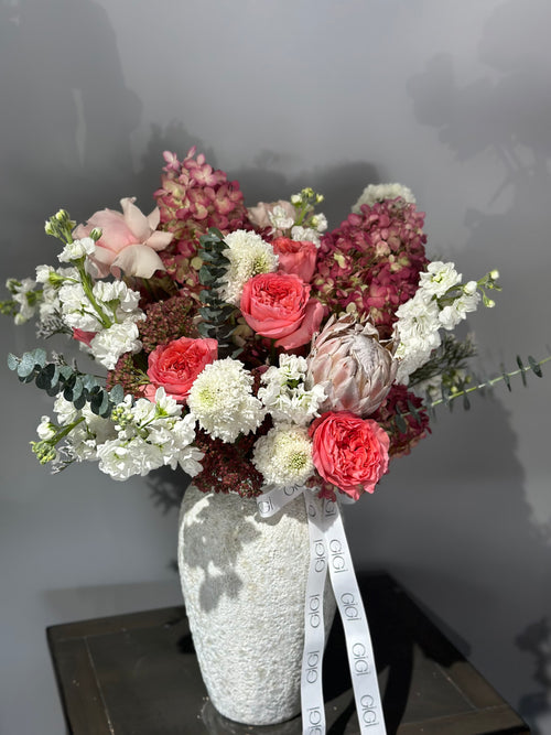 A floral bouquet featuring a variety of flowers including pink roses, white lilies, and other mixed blooms, arranged in a white vase with a lace cover.