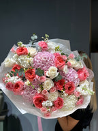 A bouquet of flowers featuring a mix of pink and white flowers, including roses and hydrangeas, wrapped in white paper.