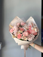 A bouquet of nude-toned roses, carnations, and ranunculus wrapped in a clear paper with a pink ribbon.