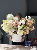 A floral arrangement featuring a variety of flowers in shades of white, beige, and brown, presented in a white cylindrical container, with a person partially visible in the background.