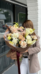 A person holding a bouquet of flowers with a variety of colors, predominantly in shades of pink and white, wrapped in a brown paper wrap with a red ribbon.