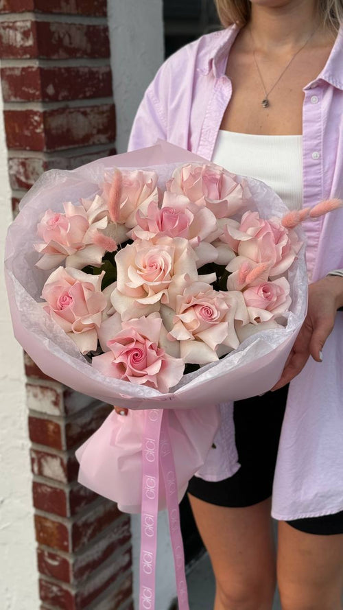 A person holding a bouquet of pink roses, the flowers are arranged in a circular shape with the stems facing upwards, wrapped with a pink and white paper.