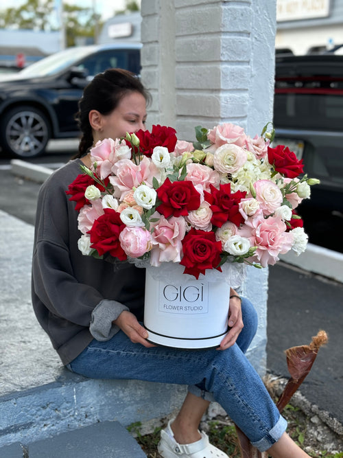 A person sitting on a curb holding a round white box of flowers with a variety of red, pink, and white flowers.