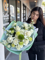 A person holding a bouquet of flowers with white roses and green foliage, wrapped in a light blue and white checkered paper.