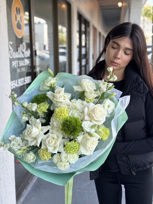 A person holding a bouquet of flowers with white roses and green foliage, wrapped in a light blue and white checkered paper.