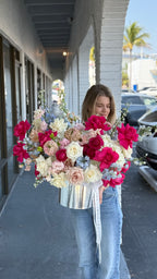 A person holding a large floral arrangement with a variety of flowers including roses in colors pink, red, white, and blue, with green leaves and stems.