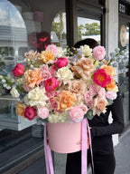 A floral arrangement featuring a variety of flowers in pink, white, and multicolor, presented in a pink box with ribbon accents.
