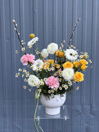A bouquet of flowers including gerberas, daisies, ranunculus, carnations, and spray roses arranged in a white vase, placed against a grey background.