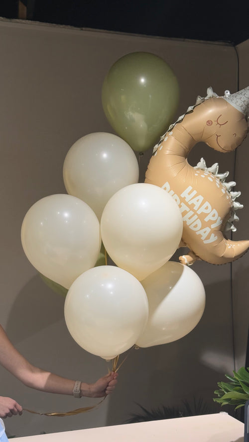 A set of balloons including three green and five white balloons, along with a dinosaur figurine with a 'Happy Birthday' inscription.