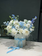 A floral arrangement featuring white and blue flowers in a round container, adorned with a blue ribbon.