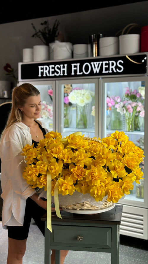 A person standing next to a basket filled with yellow roses, with a sign in the background that reads 'FRESH FLOWERS'.
