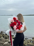 A person holding a bouquet of red and pink roses by a body of water with a cloudy sky in the background.
