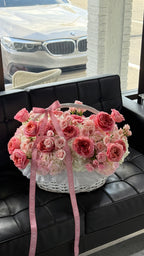 A basket of pink and white roses with hydrangeas, placed on a leather bench near a window with a car visible outside.