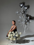 A person seated beside a large arrangement of white flowers with silver balloons in the background.