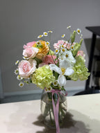 A floral bouquet featuring roses, hydrangeas, and other mixed flowers in a clear glass vase, adorned with a pink ribbon.