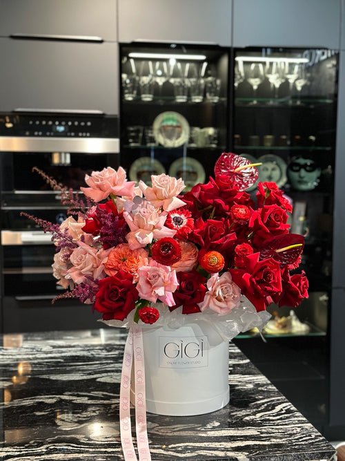 A floral arrangement featuring a mix of pink and red roses, with matthiola flowers, presented in a white container with a Gucci brand tag, displayed on a kitchen counter.