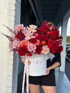 A person holding a white flower box filled with an assortment of flowers including red and pink roses, and other greenery.