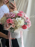A floral arrangement featuring a mix of pink and white roses, rice, and carnations, presented in a flower box with a ribbon.