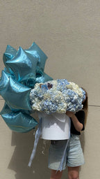 A person holding a blue hydrangea flower arrangement in a cloud-shaped box against a neutral background.