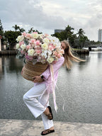 A person standing by a large basket filled with pink roses, lush hydrangeas, and orchids, overlooking a body of water.