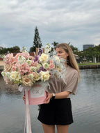 A person holding a pink floral arrangement box with a clear front, containing a variety of flowers including pink and white roses and other plants, with a background that includes a body of water and a building.