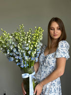 A person holding a vase with blue delphinium flowers and a mix of other flowers, with a floral pattern on the dress in the background.