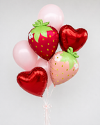 A cluster of foil balloons including two strawberry-shaped balloons with green tops, one red heart balloon, and three pale pink round balloons, tied together with a light pink ribbon against a light gray background.