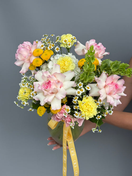 A vibrant floral bouquet held by hands against a solid gray background. The bouquet features large pink and white roses, yellow carnations, white daisies, and green foliage, tied with a yellow ribbon printed with the brand name 'GiGi Flowers'