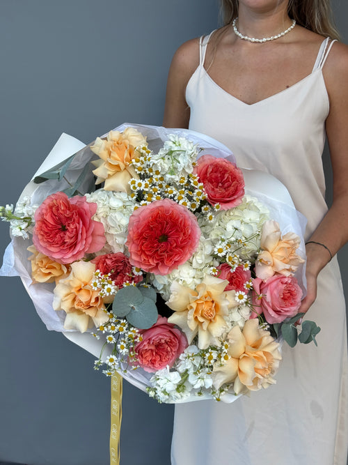 A woman in a white satin dress holds a large, vibrant bouquet of fresh flowers, including pink and peach roses, white daisies, and green eucalyptus, against a solid dark gray background.