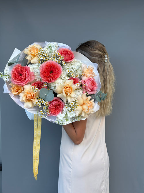 A woman in a white dress holds a large, vibrant bouquet of flowers in front of her face, showing the back of her head and the side of her body against a solid dark gray wall.