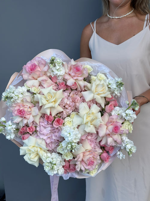 A large heart-shaped bouquet of pink, cream, and white flowers held by a woman wearing a white spaghetti-strap dress against a dark gray wall.