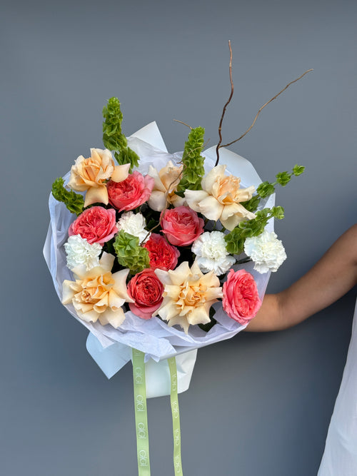 A hand holding a wrapped bouquet of mixed flowers, including peach, coral, red, and white blooms with green foliage, against a plain gray background.