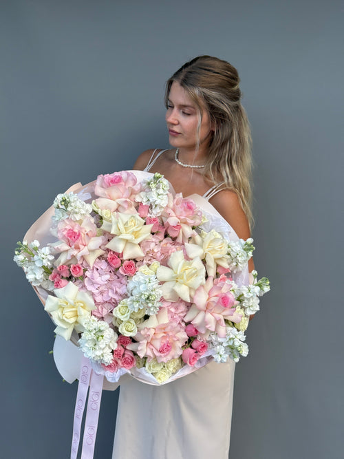 A woman in a white dress holds a large bouquet of pink, cream, and white flowers against a plain gray background.