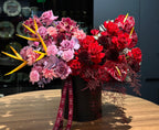 A vibrant floral arrangement featuring a mix of red and pink roses, purple flowers, and yellow tulips, presented in a black vase with a decorative ribbon.